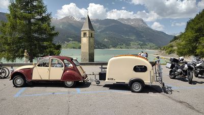Citoren 2CV mit einem farblich passenden KLEOX festival Mini-Campinganhänger mit Holzaufbau vor dem versunkenen Kirchturm von Alt-Graun am Reschensee in Südtirol