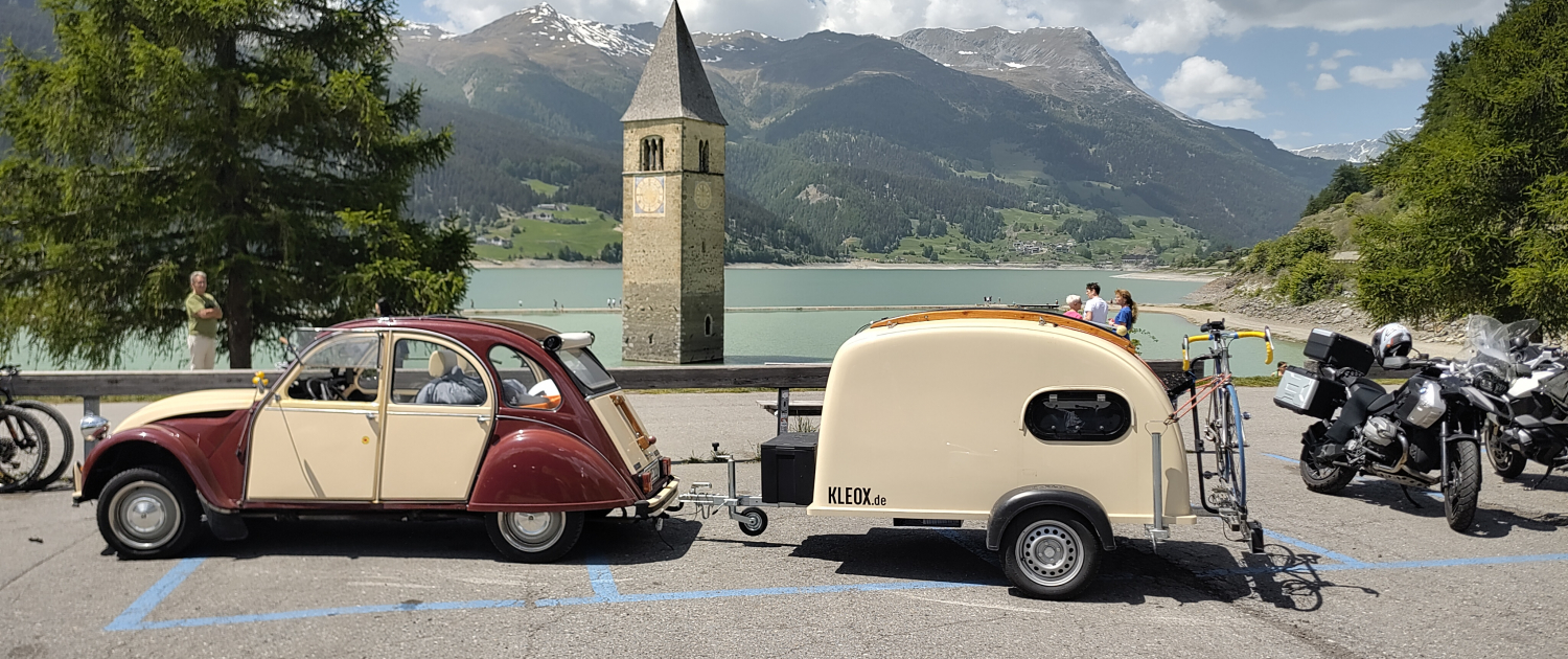 Citoren 2CV mit einem farblich passenden KLEOX festival Mini-Campinganhänger mit Holzaufbau vor dem versunkenen Kirchturm von Alt-Graun am Reschensee in Südtirol